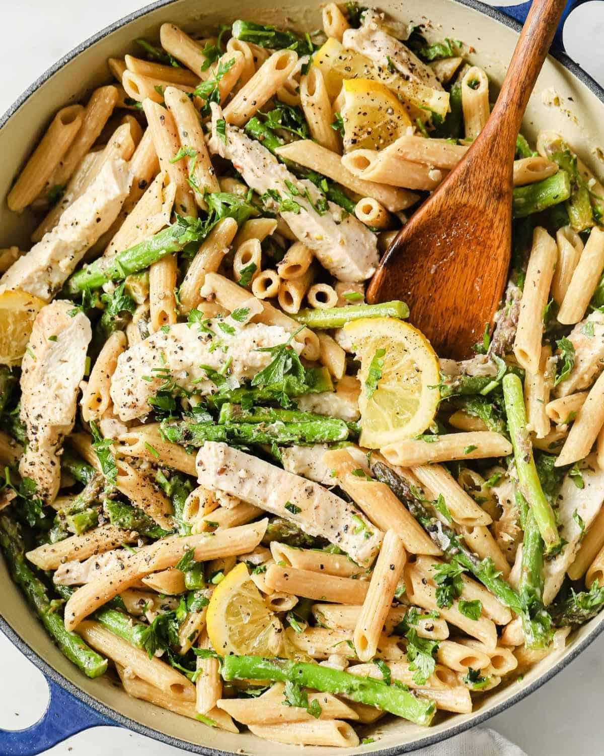 a skillet with asparagus chicken pasta and a wooden spoon