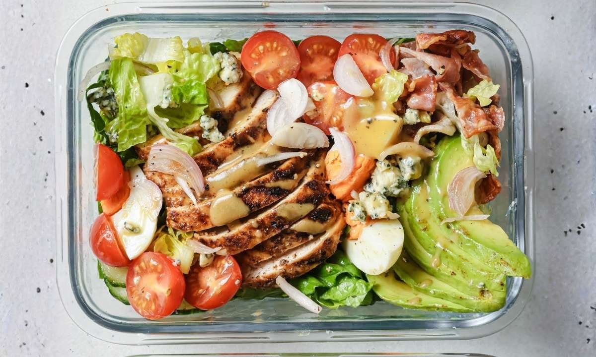 cobb chicken salad with avocado, hard-boiled eggs, tomatoes, cucumber, lettuce, and onions in a glass container