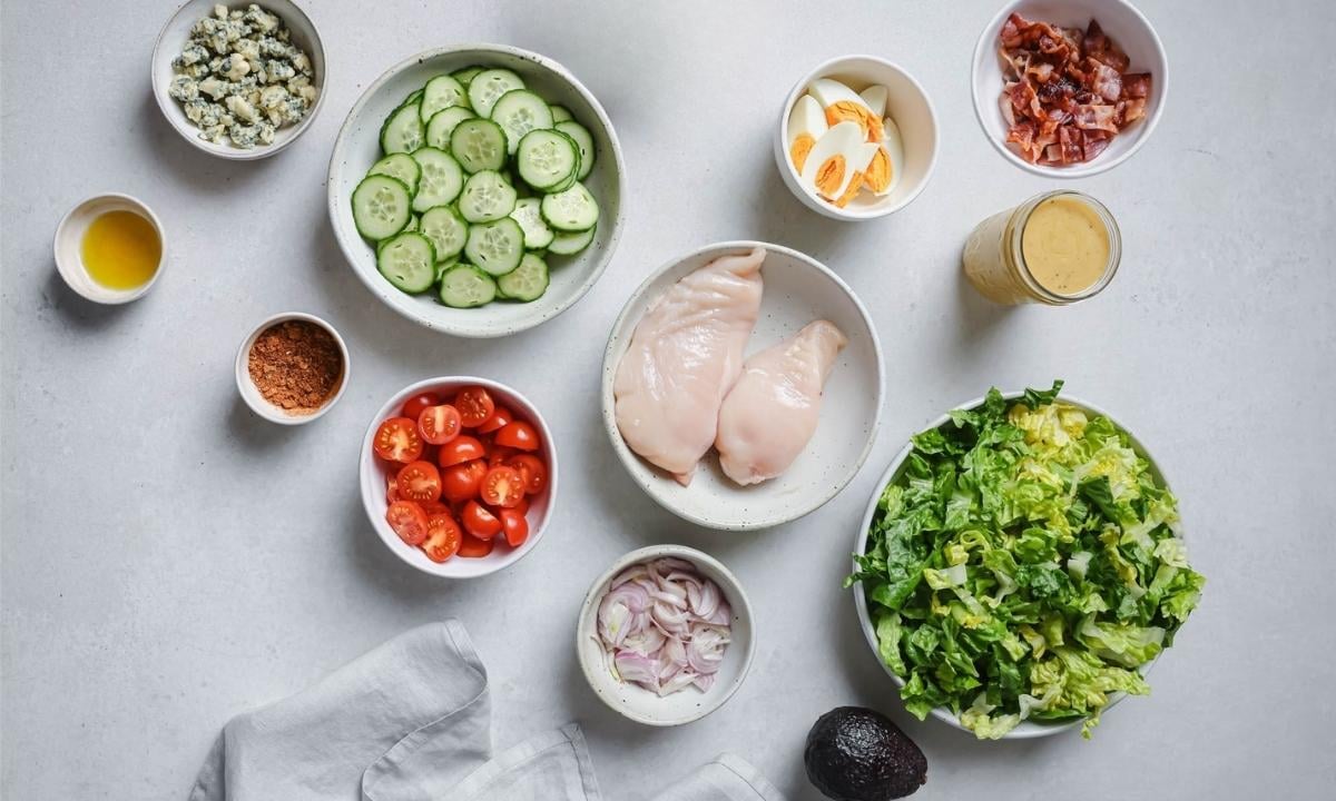 ingredients for Cobb salad in bowls: lettuce, avocado, onion, tomato, two chicken breasts, cucumber, hard-boiled egg, bacon, cheese, seasonings and a jar of dressing