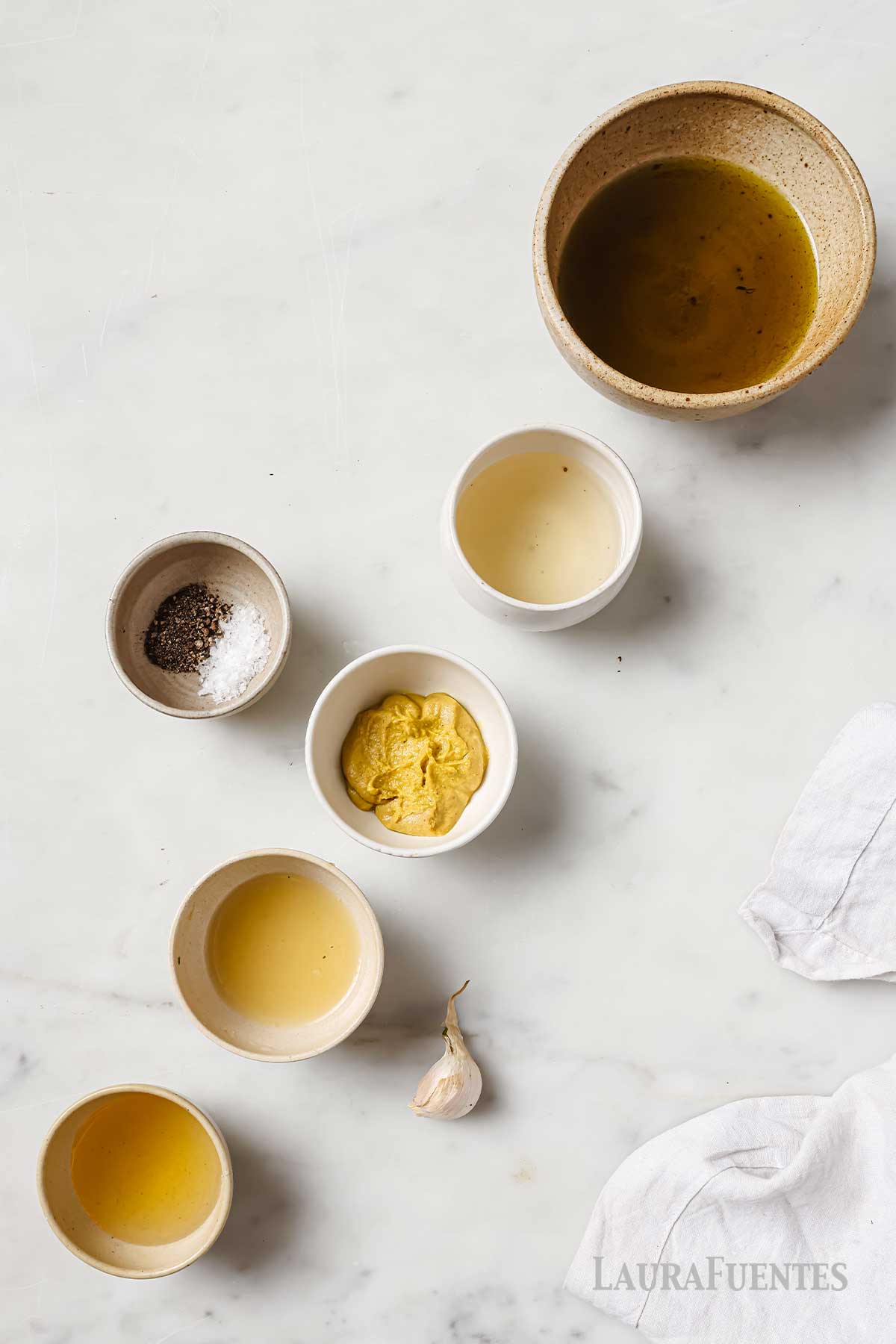 six bowls with mustard, white wine vinegar, lemon juice, honey, oil, salt and pepper, and a garlic clove next to them.