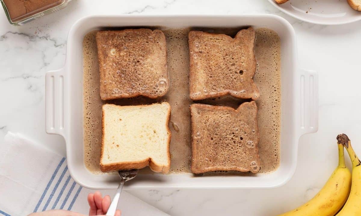 three bread slices soaked in egg banana mixture in a baking dish, and one slice being flipped