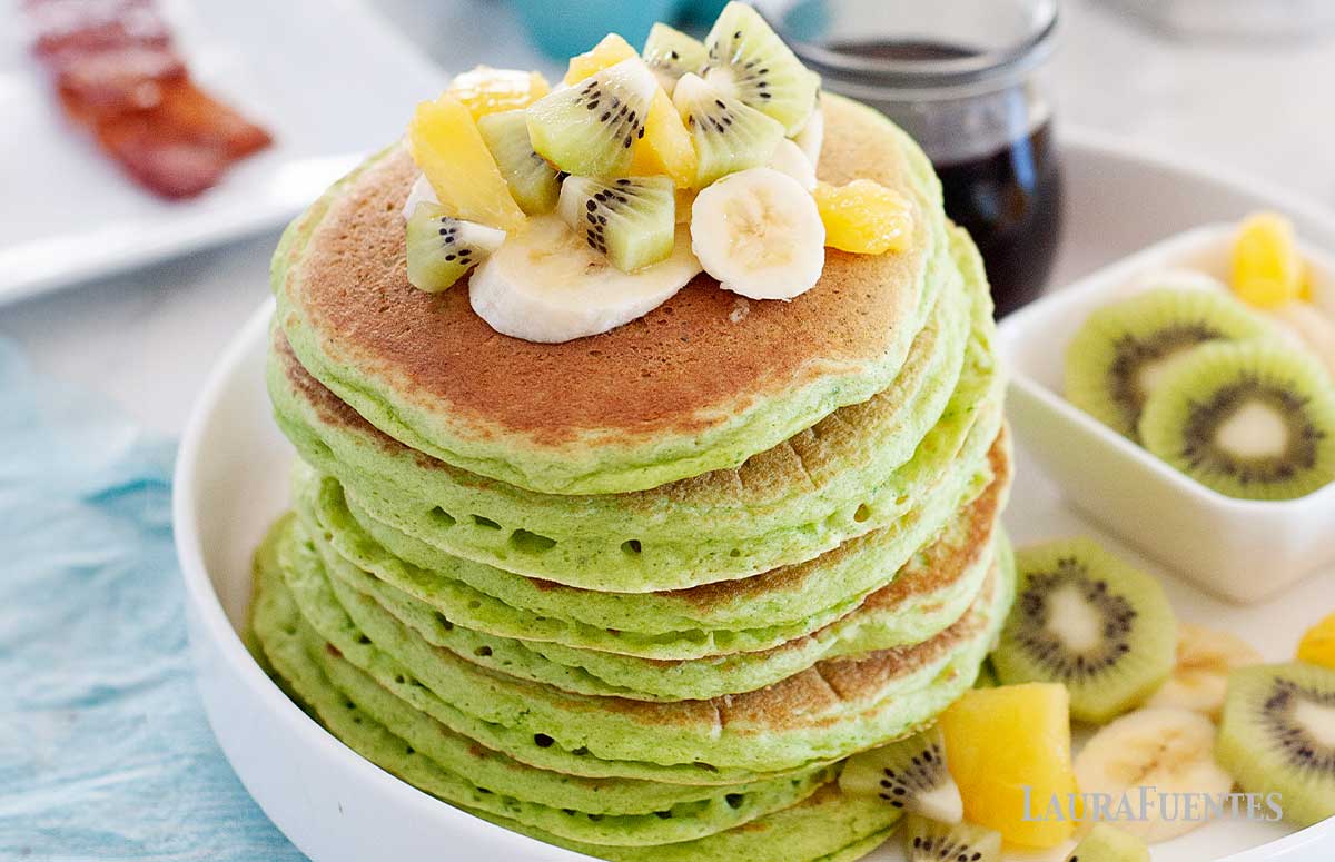 green pancakes in a large stack topped with fruit on a tray with a small cup of syrup behind.