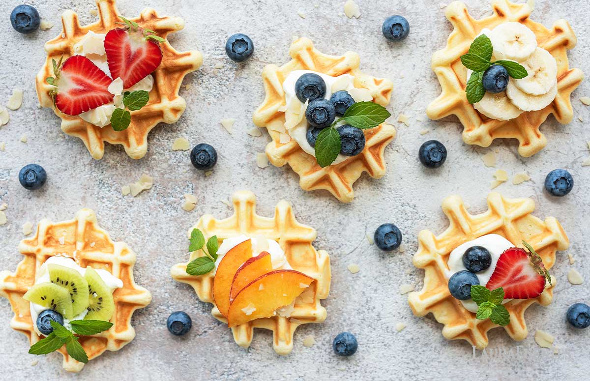overhead view of six mini waffles with different toppings on top: strawberry slices, blueberries, kiwi slices, peach slices and mint leaves