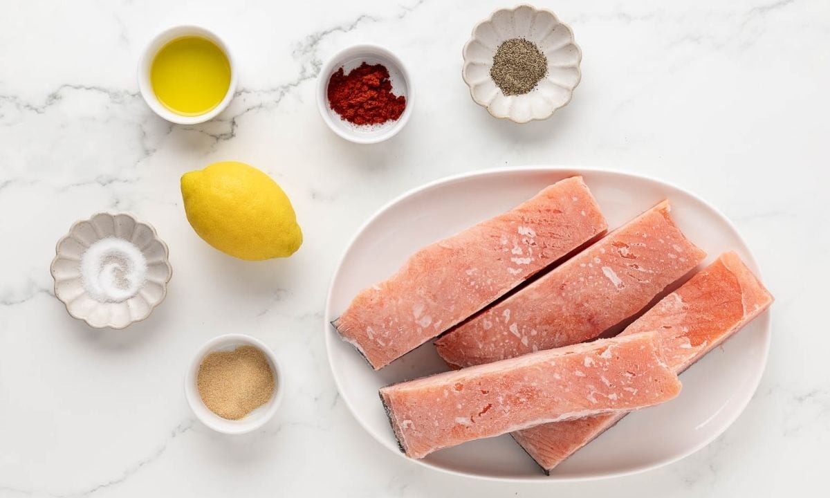 overhead view of ingredients to make delicious frozen salmon in the oven. frozen salmon pieces on a plate plus seasonings and a lemon on the counter