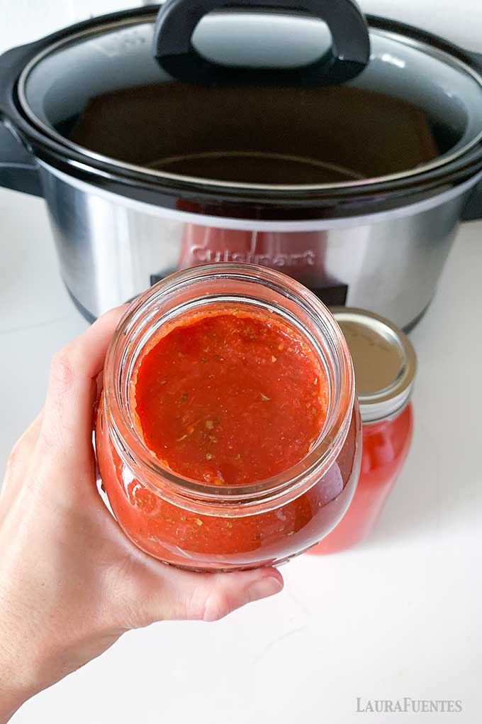 woman holding a jar of tomato sauce in her hand with a slow cooker in the background