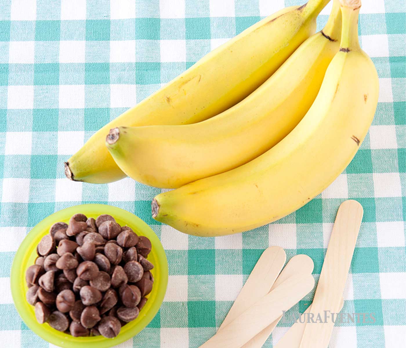 bunch of bananas on a blue tablecloth with a small green bowl of chocolate chips