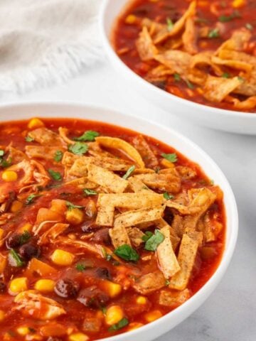 bowl filled with chicken tortilla soup (red tomato base, corn, black beans) topped with tortilla strips at bottom front of frame, same soup in a bowl in background