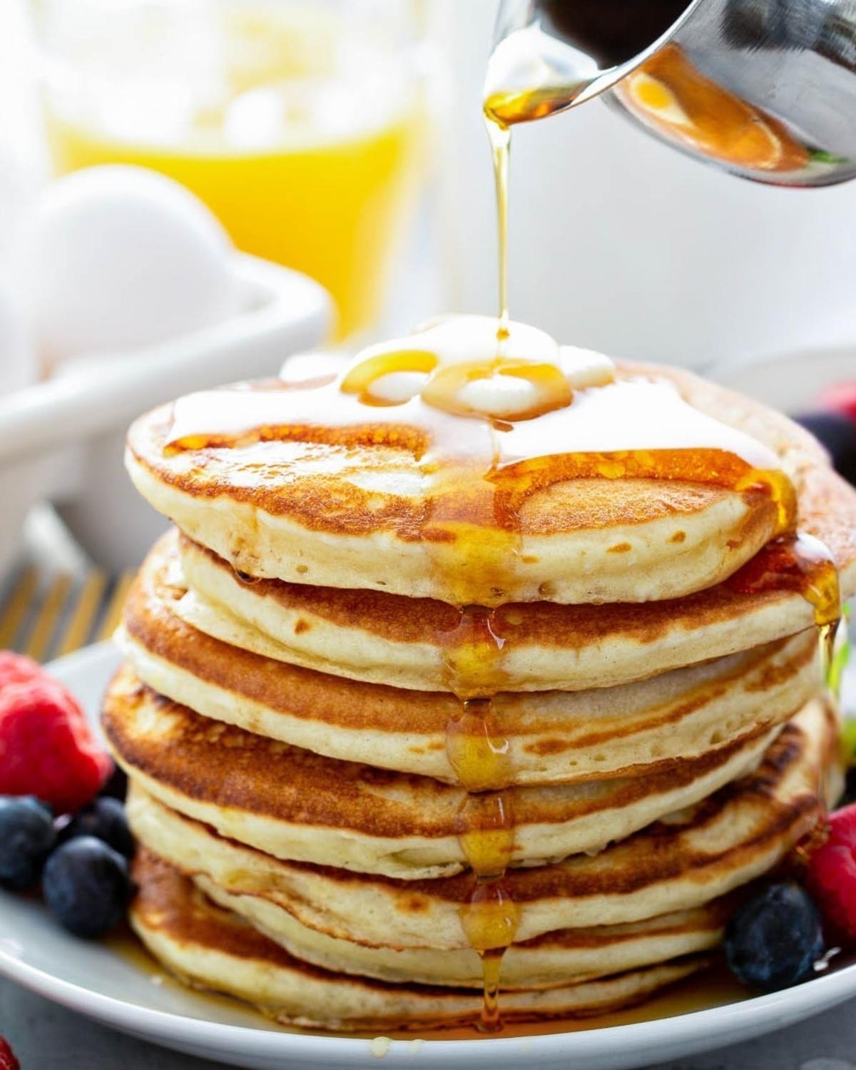 a fluffy stack of golden yeast pancakes on a plate topped with butter and syrup poured over the top and berries on the side
