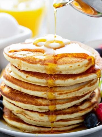 a fluffy stack of golden yeast pancakes on a plate topped with butter and syrup poured over the top and berries on the side
