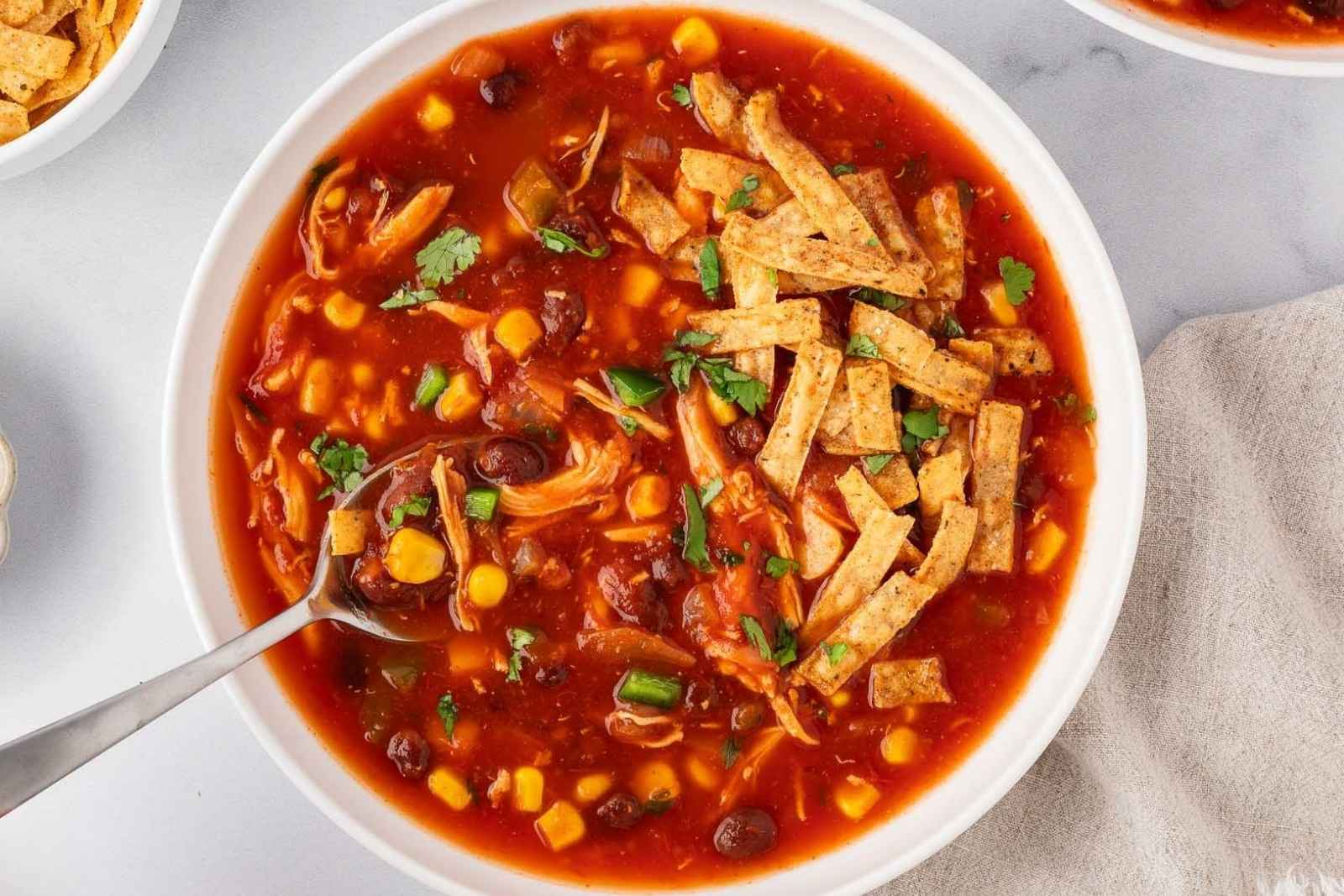 bowl filled with chicken tortilla soup (red tomato base, corn, black beans) topped with tortilla strips and a spoon filled with the soup