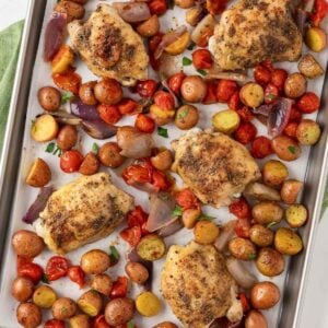 overhead view baked chicken thighs on a sheet pan with tomatoes, baby potatoes, and onions