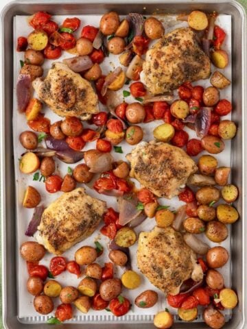 overhead view baked chicken thighs on a sheet pan with tomatoes, baby potatoes, and onions