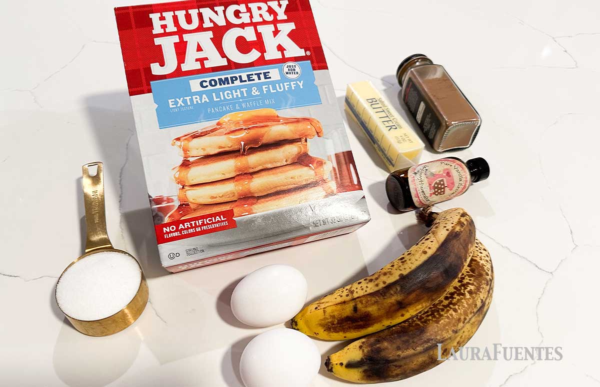 a box of pancake mix, two eggs, two bananas, a bottle of vanilla extract, a stick of butter, a jar with cinnamon, and a spoon with sugar
