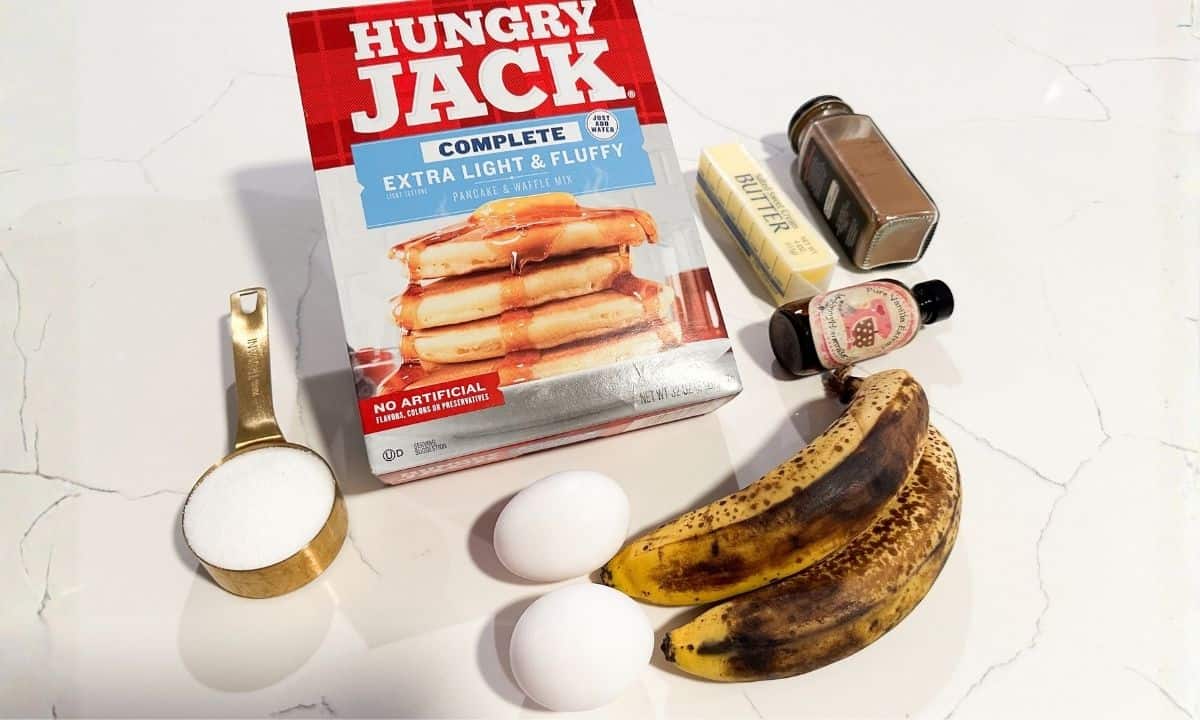 a box of pancake mix, two eggs, two bananas, a bottle of vanilla extract, a stick of butter, a jar with cinnamon, and a spoon with sugar