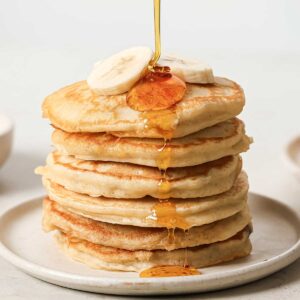 a stack of banana pancakes on a plate with sliced bananas on top and syrup being drizzled over them
