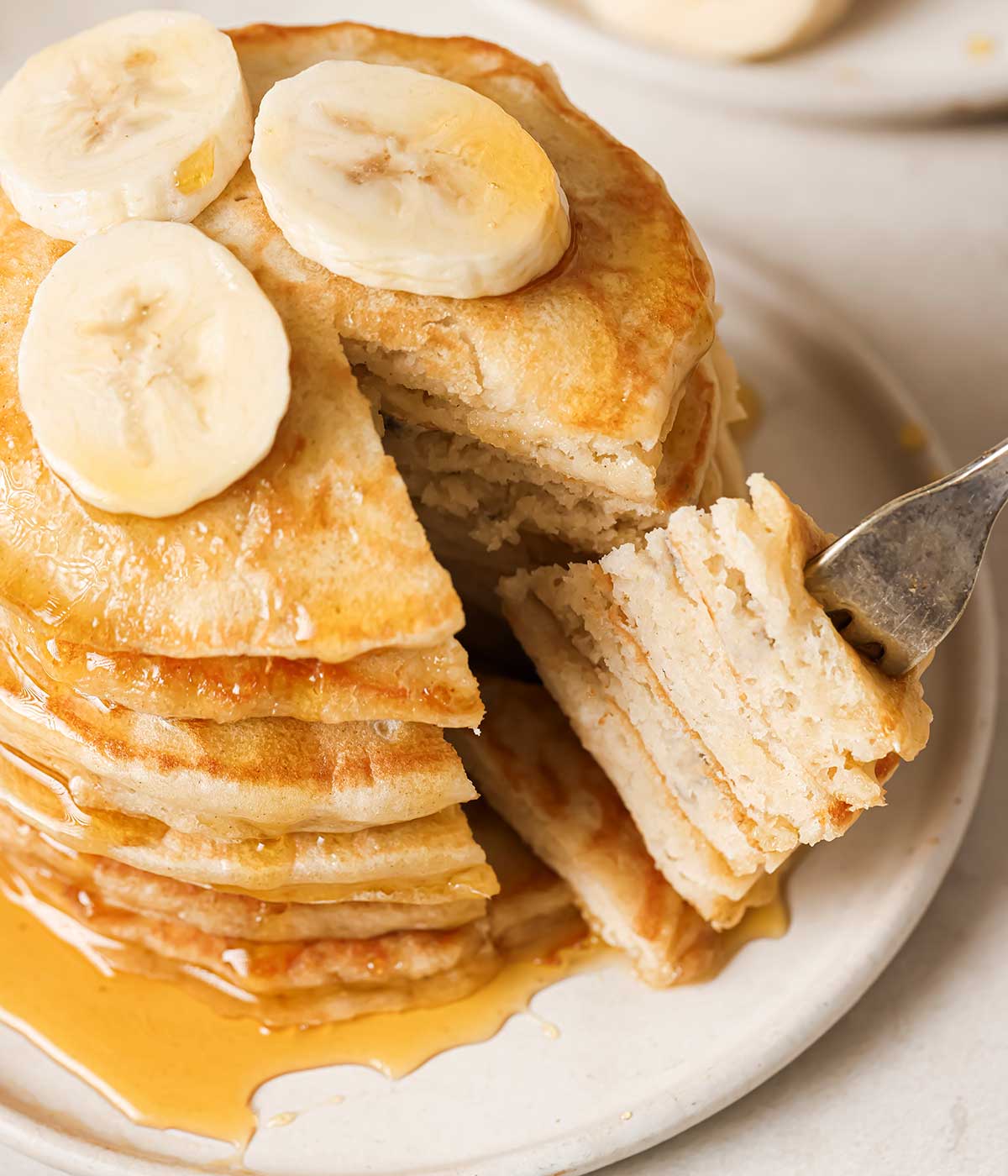 a stack of banana pancakes on a plate with sliced bananas on top and syrup, where the pancakes have been sliced and are poked on a fork to show the fluffy texture