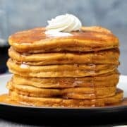 tall fluffy stack of pumpkin pancakes made with cottage cheese on a plate topped with a little whipped cream and pancake syrup