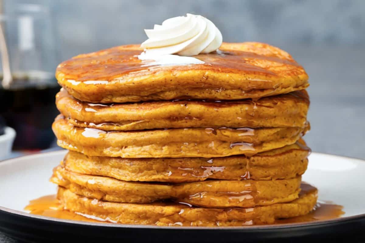 tall fluffy stack of pumpkin pancakes made with cottage cheese on a plate topped with a little whipped cream and pancake syrup
