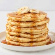 stack of banana protein powder pancakes on a plate topped with bananas and lightly drizzled with syrup
