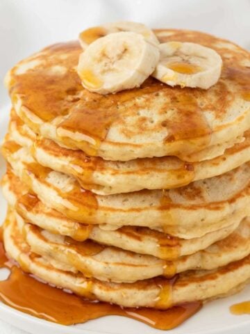 stack of banana protein powder pancakes on a plate topped with bananas and lightly drizzled with syrup