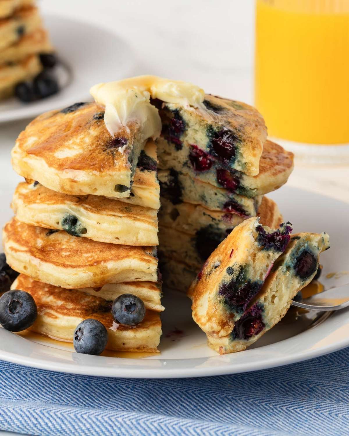 Stack of blueberry buttermilk pancakes on a plate topped with butter, syrup, and some blueberries on the plate and a piece has been sliced to show their fluffy texture