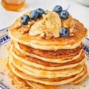 stack of banana nut pancakes on a plate topped with sliced bananas, chopped walnuts, blueberries, and pancake syrup