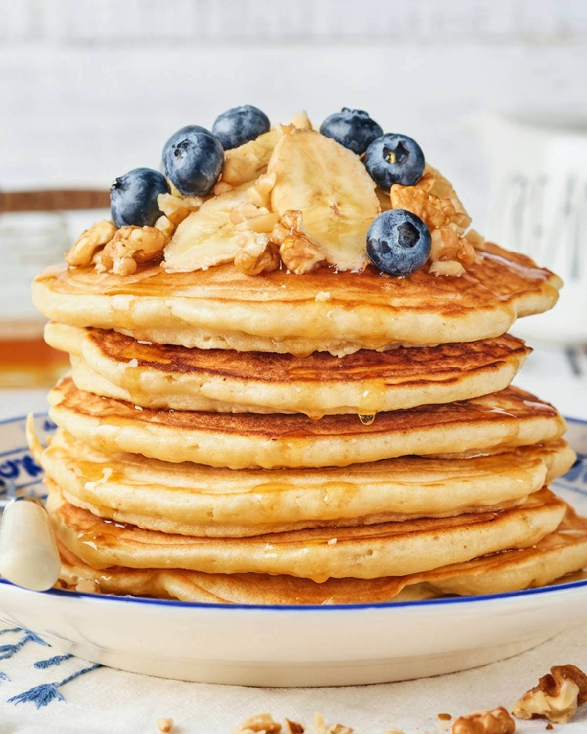 stack of banana nut pancakes on a plate topped with sliced bananas, chopped walnuts, blueberries, and pancake syrup