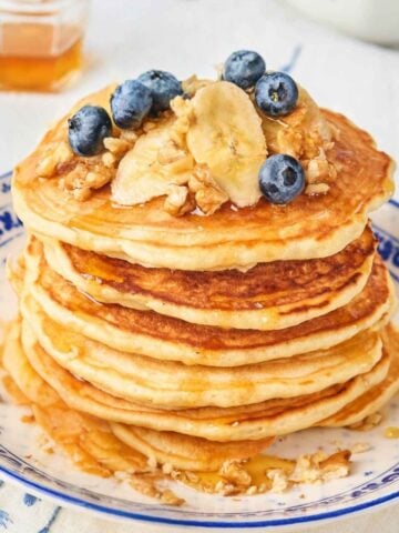 stack of banana nut pancakes on a plate topped with sliced bananas, chopped walnuts, blueberries, and pancake syrup