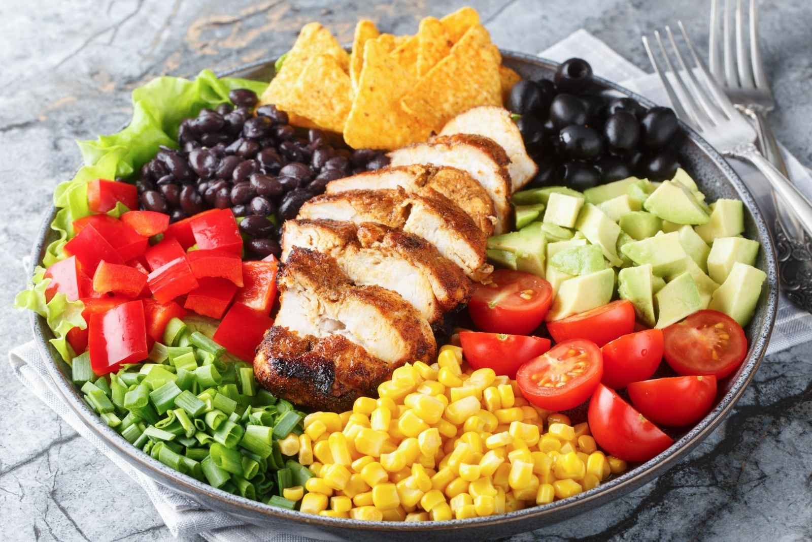 a large bowl of Mexican salad made with corn, tomatoes, chicken, beans, avocado and chips