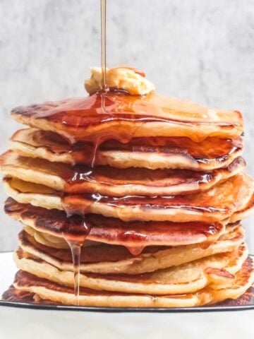 large stack of pancakes topped with butter with syrup being poured over the top