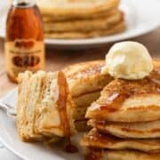 stack of homemade cracker barrel pancakes on a plate, sliced and a bite is on a fork, topped with whipped butter and syrup with a syrup bottle on background