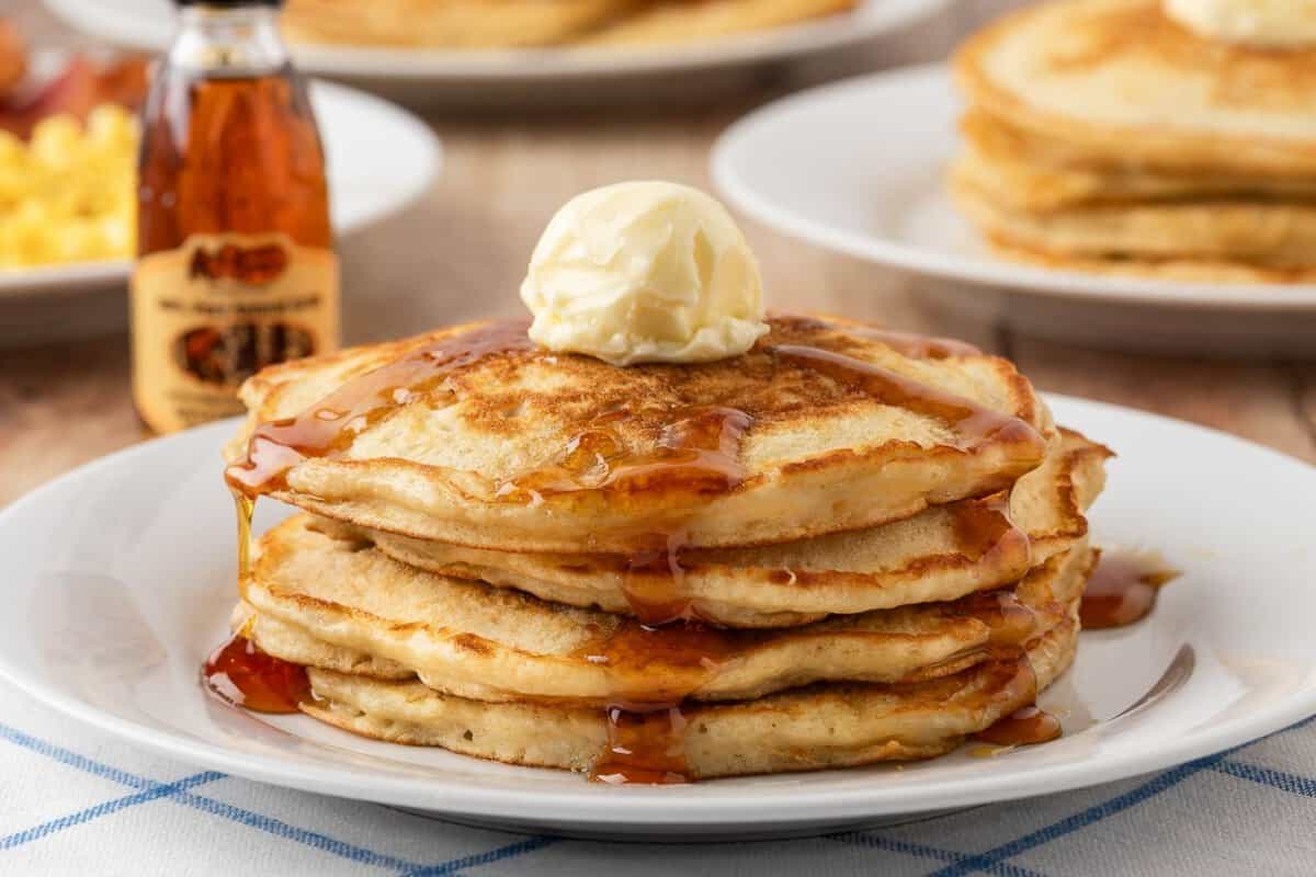 stack of homemade cracker barrel pancakes on a plate, topped with whipped butter and syrup with a syrup bottle on background