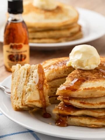 stack of homemade cracker barrel pancakes on a plate, sliced and a bite is on a fork, topped with whipped butter and syrup with a syrup bottle on background