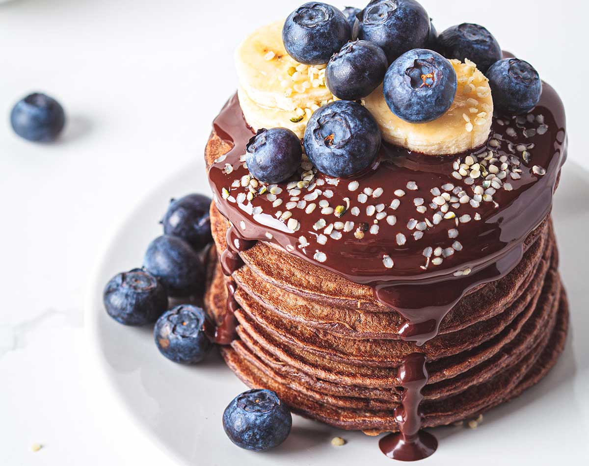 overhead view of a stack of chocolate banana pancakes with blueberries and chocolate syrup on a plate