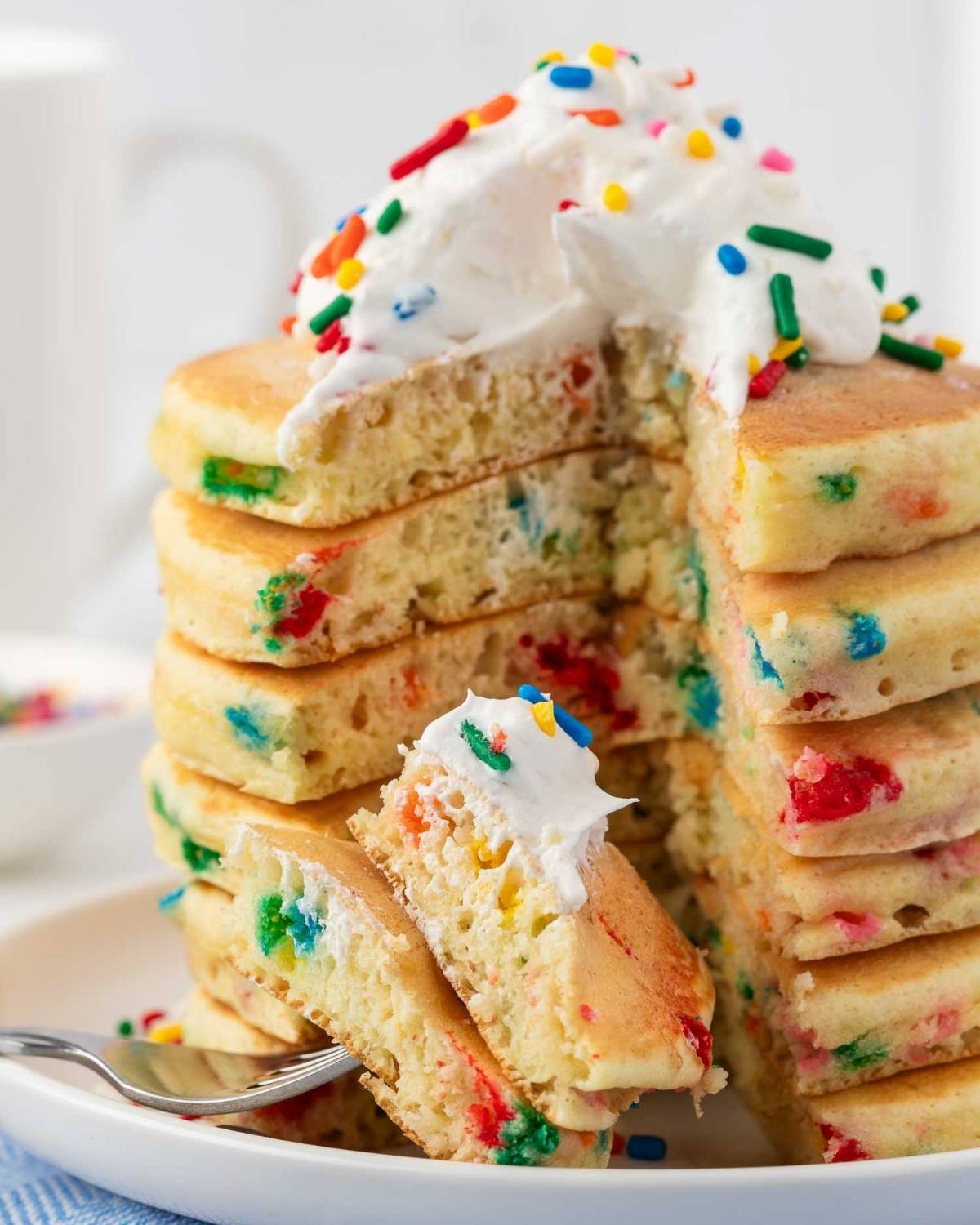 stack of birthday cake pancakes sliced to show the funfetti sprinkles inside and fluffy texture. they're topped with whipped cream and sprinkles