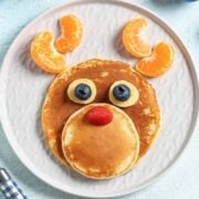 reindeer pancake on a plate made with fluffy pancakes, banana slices, and blueberries as eyes with orange slices as antlers.