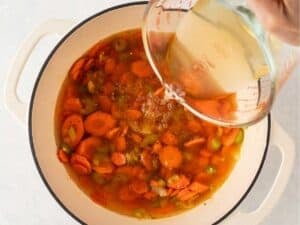 Chicken broth being poured into a pot of sautéed carrots, celery, onions, and garlic for chicken noodle soup.