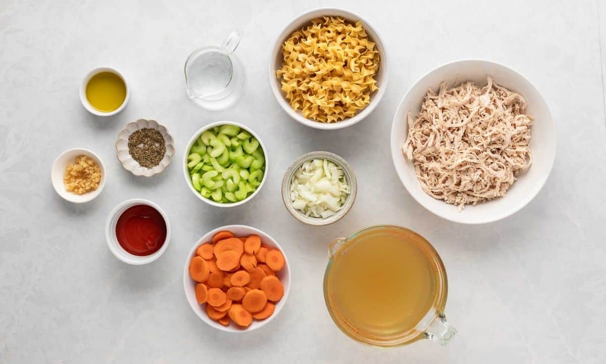 ingredients for spicy chicken noodle soup, including shredded chicken, egg noodles, chicken broth, carrots, celery, onion, garlic, sriracha, oil, water, and seasonings.