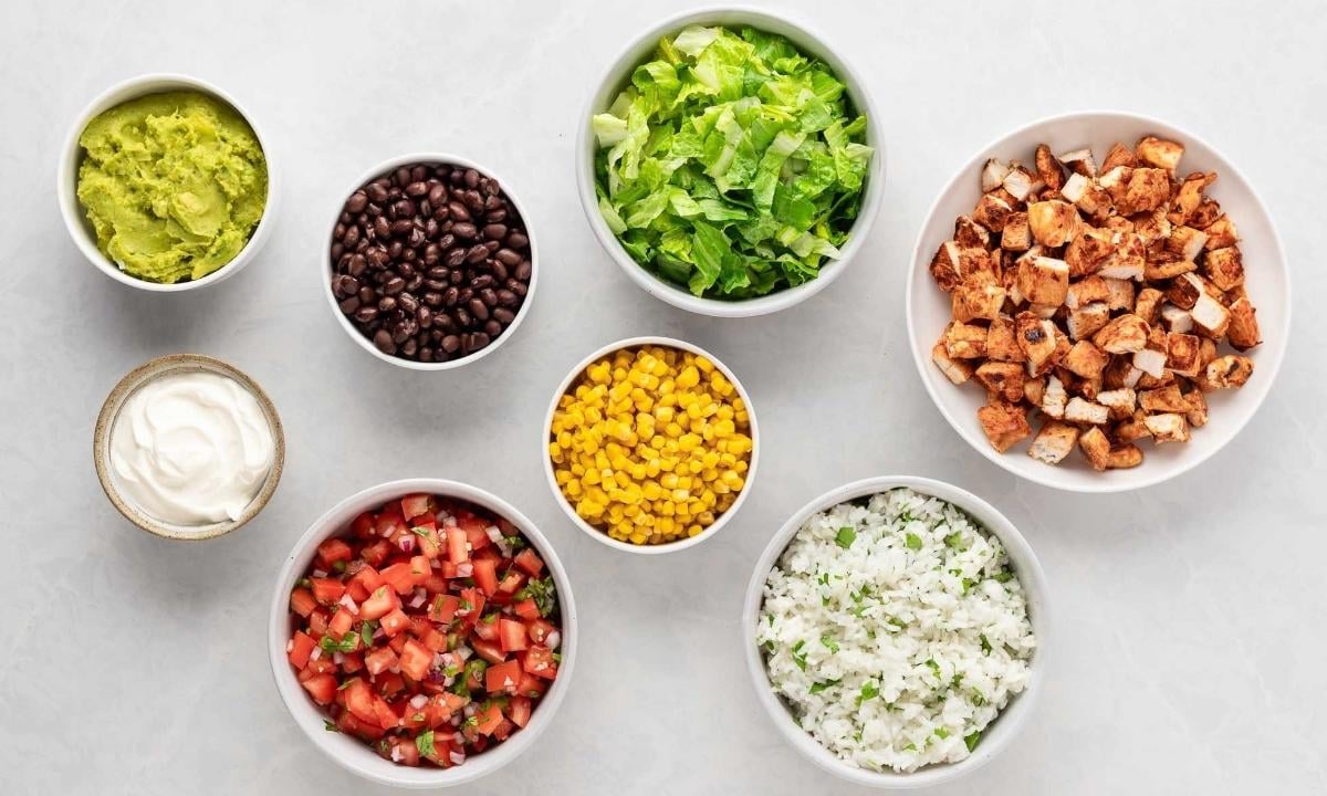 small white bowls individually filled with guacamole, sour cream, black beans, pico de gallo, corn, cilantro lime rice, cooked chicken cubes, and lettuce