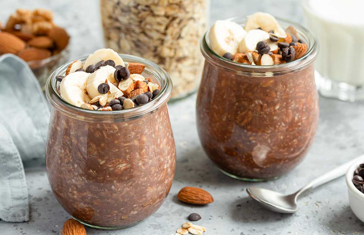 two jars of chocolate protein overnight oats topped with banana slices, chopped walnuts and chocolate chips