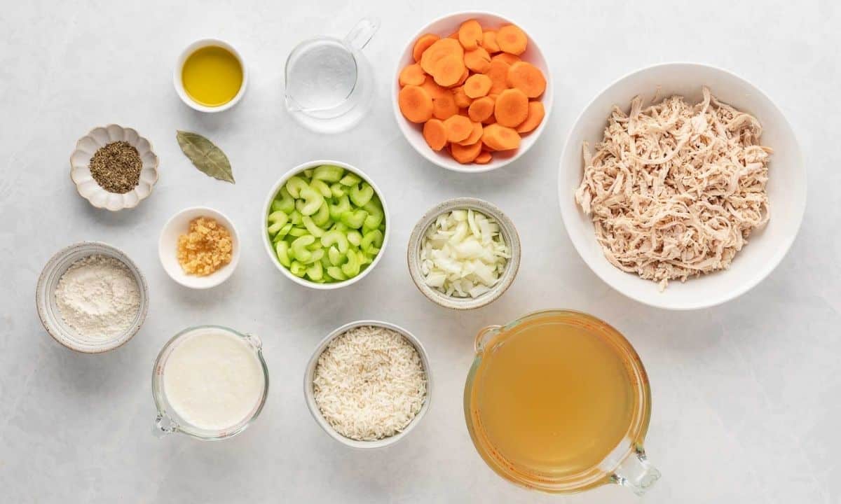 An overhead shot of ingredients for chicken rice soup, including shredded chicken, rice, chicken broth, sliced carrots, chopped celery, and onions, seasonings in separate bowls.