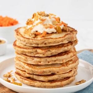 stack of carrot cake pancakes on a plate topped with a bit of cream cheese frosting, shredded carrot sprinkles, and chopped walnuts
