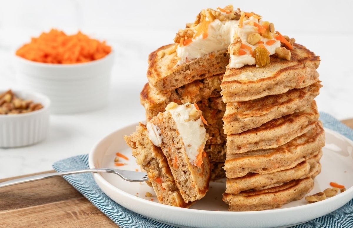 a slice poked on a fork of carrot cake pancakes. showing fluffy texture and a close up of the carrot cake bits inside. Topped with cream cheese frosting