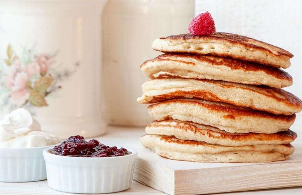 fluffy stack of sour cream pancakes with a raspberry on top stacked on a wooden board. small dish of sour cream next to board and another small dish with jam