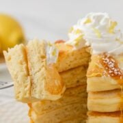 fluffy stack of lemon ricotta pancakes, two pieces on a fork to show fluffy texture, topped with whipped cream and shaved lemon with a drizzle of pancake syrup
