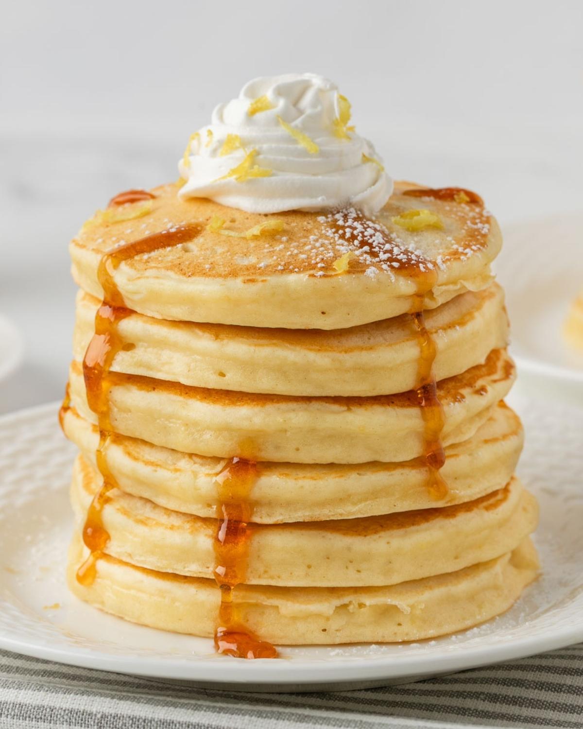 fluffy stack of lemon ricotta pancakes topped with whipped cream and shaved lemon with a drizzle of pancake syrup