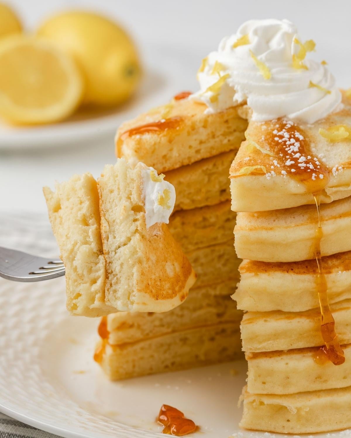 fluffy stack of lemon ricotta pancakes, two pieces on a fork to show fluffy texture, topped with whipped cream and shaved lemon with a drizzle of pancake syrup