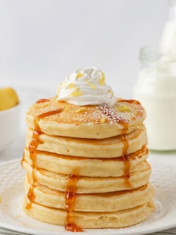 fluffy stack of lemon ricotta pancakes topped with whipped cream and shaved lemon with a drizzle of pancake syrup