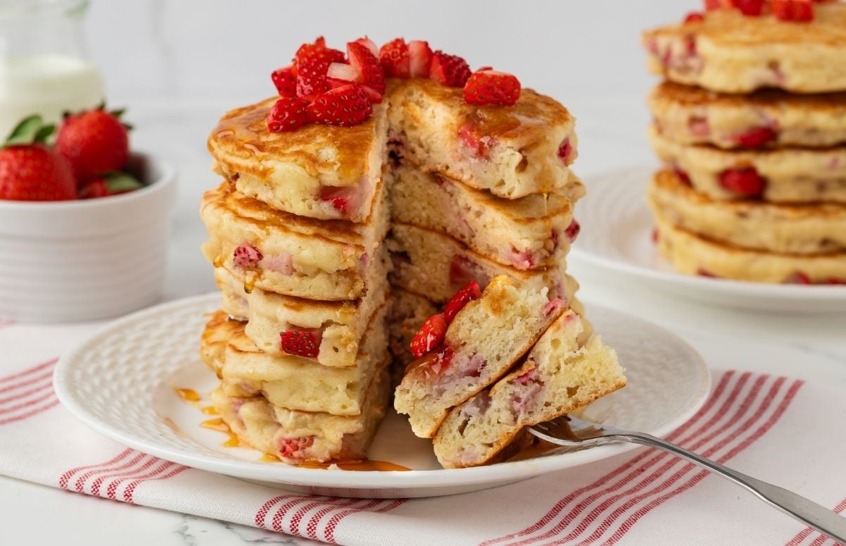 Fluffy stack of strawberry pancakes on a white plate. A slice has been cut and is poked on a fork to show the fluffy texture. The image has more pancakes in the background and more strawberries.