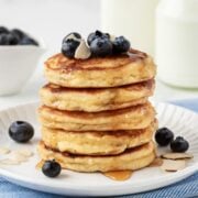 Front view of a fluffy stack of almond flour pancakes on a plate topped with blueberries and some almond slivers with a little maple syrup drizzled on top.